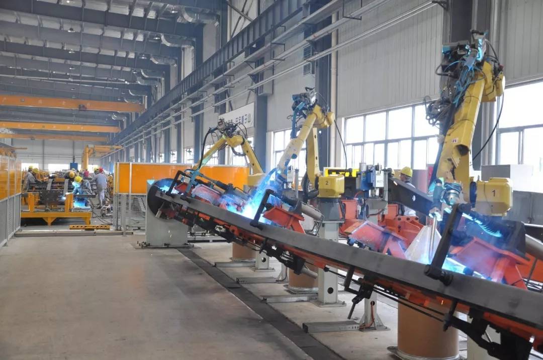 #image_title Robotic welding line in a spacious factory with yellow arms welding metal on a tilted conveyor; workers are seen in the background.