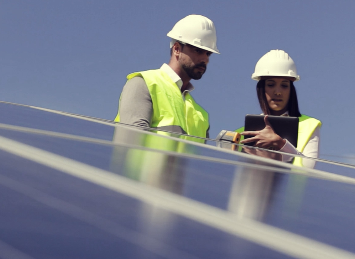 #image_title Two construction workers in hard hats and high-visibility vests inspect a row of solar panels, one holding a tablet and the other monitoring with a device.